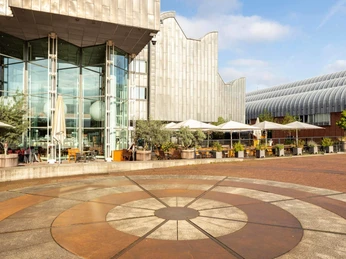 MAALOT Das Bild zeigt eine moderne Architektur mit großflächigen Glasfassaden und einem weitläufigen Platz davor. Im Vordergrund ist ein kreisförmiges Bodendesign zu erkennen, während Tische und Stühle unter weißen Sonnenschirmen auf eine einladende Atmosphäre hinweisen.The picture shows modern architecture with large glass façades and a spacious square in front of it. A circular floor design can be seen in the foreground, while tables and chairs under white parasols suggest an inviting atmosphere.