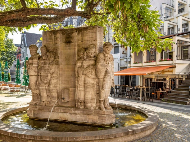 Ostermann-Brunnen Das Bild zeigt den Ostermann-Brunnen in Köln an einem sonnigen Tag. Mehrere Statuen umringen den rechteckigen Brunnen, aus dem Wasser sprudelt. Im Hintergrund sind ein Straßencafé und mehrere Gebäude mit Balkonen zu sehen, die von Bäumen beschattet werden.