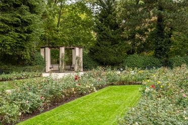 Rosengarten im Fort X Der Rosengarten am Fort X in Köln erstreckt sich unter hohen Bäumen und bietet einen Pavillon, umgeben von bunten Rosenblüten und gepflegten Rasenflächen.