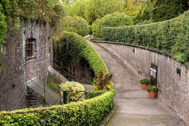 Rose Garden at Fort X The picture shows a picturesque path lined with hedges that leads through the historic rose garden at Fort X in Cologne. Old stone facades and a staircase flank the peaceful path. The scene captivates with lush greenery and an atmosphere of tranquillity.