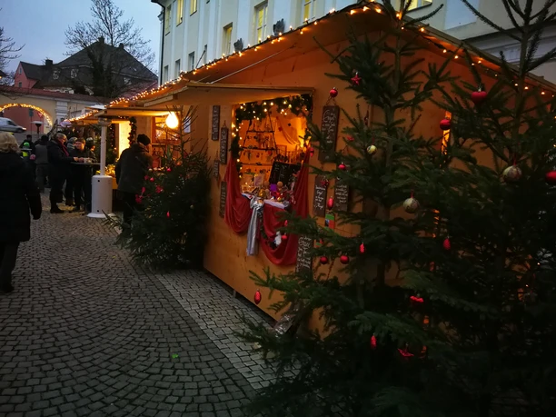 Ein festlich beleuchteter Weihnachtsmarktstand mit Dekorationen und Tannen befindet sich in einer charmanten Altstadtgasse in Günzburg, umgeben von winterlich gekleideten Menschen.