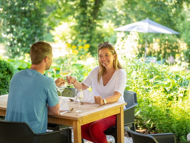 lage-hoerster-egge-haus-berkenkamp-teutoburger-wald-tourismus-d-ketz-034-jpg.jpg Ein Paar sitzt im sommerlichen Garten an einem Holztisch und stößt mit Weingläsern an.