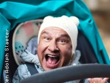 Lächelnder Mann mit weißer Mütze in blauem Kinderwagen vor verschwommenem, herbstlichem Hintergrund.