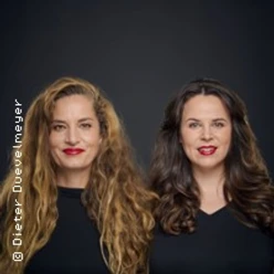 Zwei lächelnde Frauen mit offenen Haaren, beide in schwarzer Kleidung, vor einem dunklen Hintergrund.