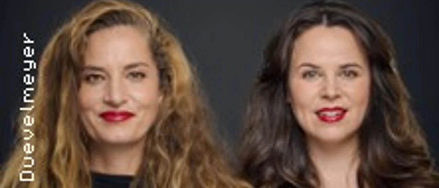 Zwei lächelnde Frauen mit offenen Haaren, beide in schwarzer Kleidung, vor einem dunklen Hintergrund.