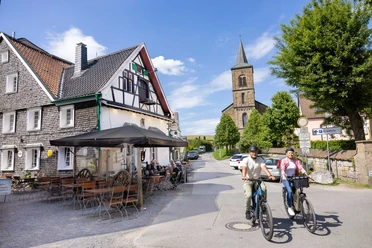 In Wülfrath-Düssel unterwegs Zwei Menschen fahren auf Fahrrädern eine Straße entlang, vorbei an Fachwerkhäusern und einer Kirche in Wülfrath-Düssel.