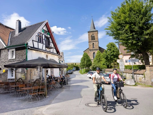 In Wülfrath-Düssel unterwegs Zwei Menschen fahren auf Fahrrädern eine Straße entlang, vorbei an Fachwerkhäusern und einer Kirche in Wülfrath-Düssel.