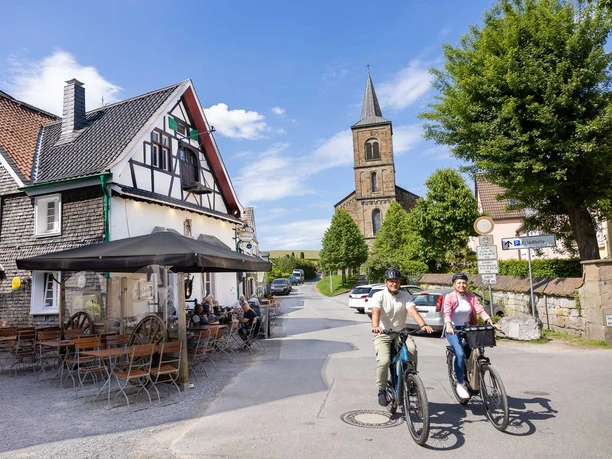 In Wülfrath-Düssel unterwegs Zwei Menschen fahren auf Fahrrädern eine Straße entlang, vorbei an Fachwerkhäusern und einer Kirche in Wülfrath-Düssel.
