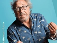 Ein älterer Mann mit Brille und wildem Haar, der ein blaues Hemd mit Tiermotiven trägt, gestikuliert.