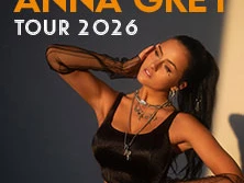 Anna Grey lehnt entspannt an einer Wand, gekleidet in ein stilvolles schwarzes Top, umrahmt von sanftem Licht.