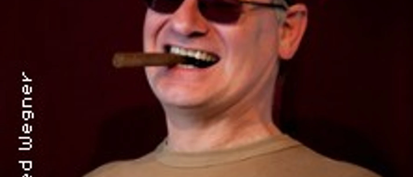 Ein älterer Mann mit roter Baskenmütze und Sonnenbrille lächelt und hält eine Zigarre im Mund.