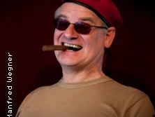 Ein älterer Mann mit roter Baskenmütze und Sonnenbrille lächelt und hält eine Zigarre im Mund.