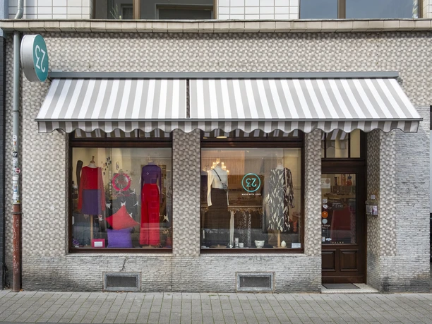 rock-it-baby Das Bild zeigt eine Ladenfassade in stilvollem Mosaikdesign. Vor den Schaufenstern hängt eine gestreifte Markise.