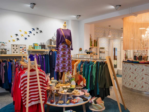 rock-it-baby Ein stilvoll eingerichtetes Modegeschäft zeigt farbenfrohe Kleidung und Accessoires auf Kleiderständern und einem Tisch.