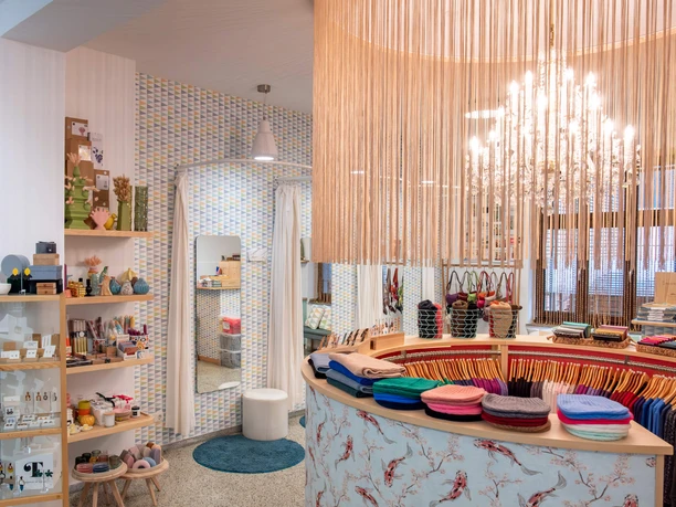 rock-it-baby Das Bild zeigt das Innere eines stilvoll eingerichteten Geschäfts mit einem modernen und einladenden Ambiente. Rechts im Vordergrund ist eine helle, halbkreisförmige Theke mit verschiedenen farbigen Tüchern sichtbar. Links sind Regale mit Kosmetikprodukten und Accessoires angeordnet. Ein Kronleuchter und helle Vorhänge schaffen ein warmes Licht.