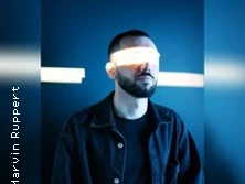 Ein Mann steht in dunkler Umgebung; Lichtstrahl verdeckt seine Augen und erzeugt dramatischen Effekt.