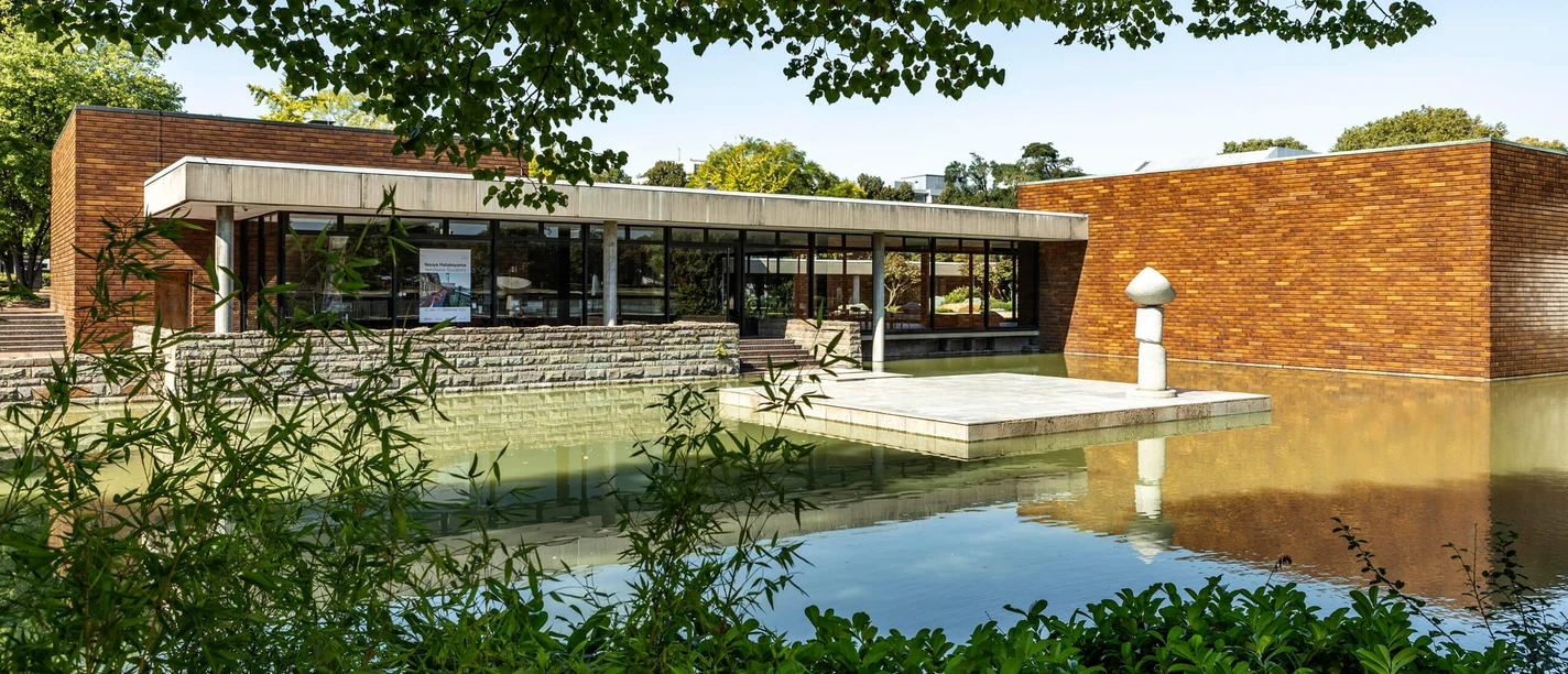Museum für Ostasiatische Kunst Museum für Ostasiatische Kunst in Köln, das von einer Wasserfläche umgeben ist. Die Architektur besteht aus modernen, geradlinigen Strukturen mit großen Fenstern und Ziegelwänden. Im Vordergrund sind grüne Pflanzen zu erkennen. Das Gebäude spiegelt sich malerisch in dem ruhigen Wasser.