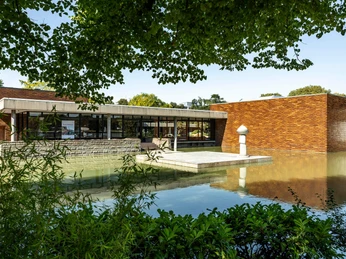 Museum für Ostasiatische Kunst Museum für Ostasiatische Kunst in Köln, das von einer Wasserfläche umgeben ist. Die Architektur besteht aus modernen, geradlinigen Strukturen mit großen Fenstern und Ziegelwänden. Im Vordergrund sind grüne Pflanzen zu erkennen. Das Gebäude spiegelt sich malerisch in dem ruhigen Wasser.Museum of East Asian Art in Cologne, which is surrounded by an expanse of water. The architecture consists of modern, linear structures with large windows and brick walls. Green plants can be seen in the foreground. The building is reflected picturesquely in the calm water.