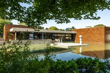 Museum of East Asian Art Museum of East Asian Art in Cologne, which is surrounded by an expanse of water. The architecture consists of modern, linear structures with large windows and brick walls. Green plants can be seen in the foreground. The building is reflected picturesquely in the calm water.