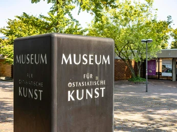 Museum für Ostasiatische Kunst Museum für Ostasiatische Kunst in Köln, moderner Bau mit dunklem Schild, grüne Bäume im Hintergrund.The image depicts the main sign of the Museum of East Asian Art in Cologne, Germany, with a modern building and lush trees in the background.
