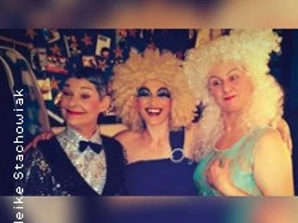 Drei Personen in extravagantem Make-up und glitzernden Kostümen vor einem bunt dekorierten Hintergrund.