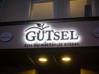 Außenansicht Restaurant Gütsel Leuchtendes "GÜTSEL"-Schild bei Nacht, darunter der Schriftzug "Echt HeimatGenuss erleben".