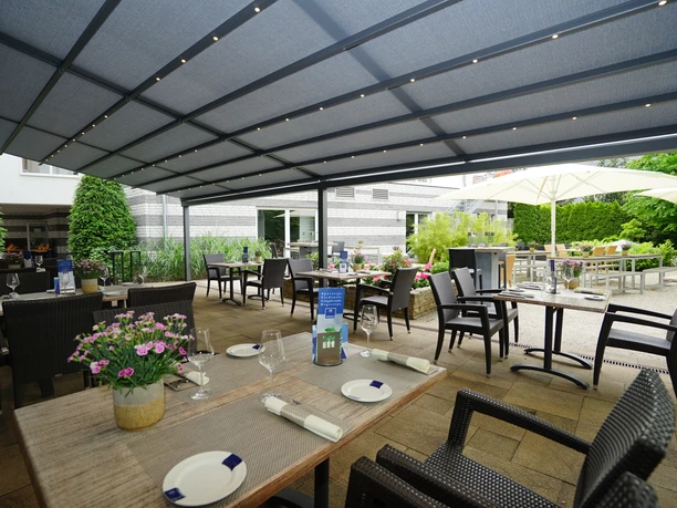 Außengastronomie im Gütesl Überdachte Außenterrasse eines Restaurants mit gedeckten Tischen, grüner Bepflanzung und Sonnenliegen.