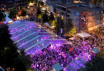 WMC Kerkade (c) WMC.jpg Menschenmenge unter einem transparenten Zelt, das in violettem Licht erstrahlt. Im Hintergrund stehen Gebäude und Bäume.