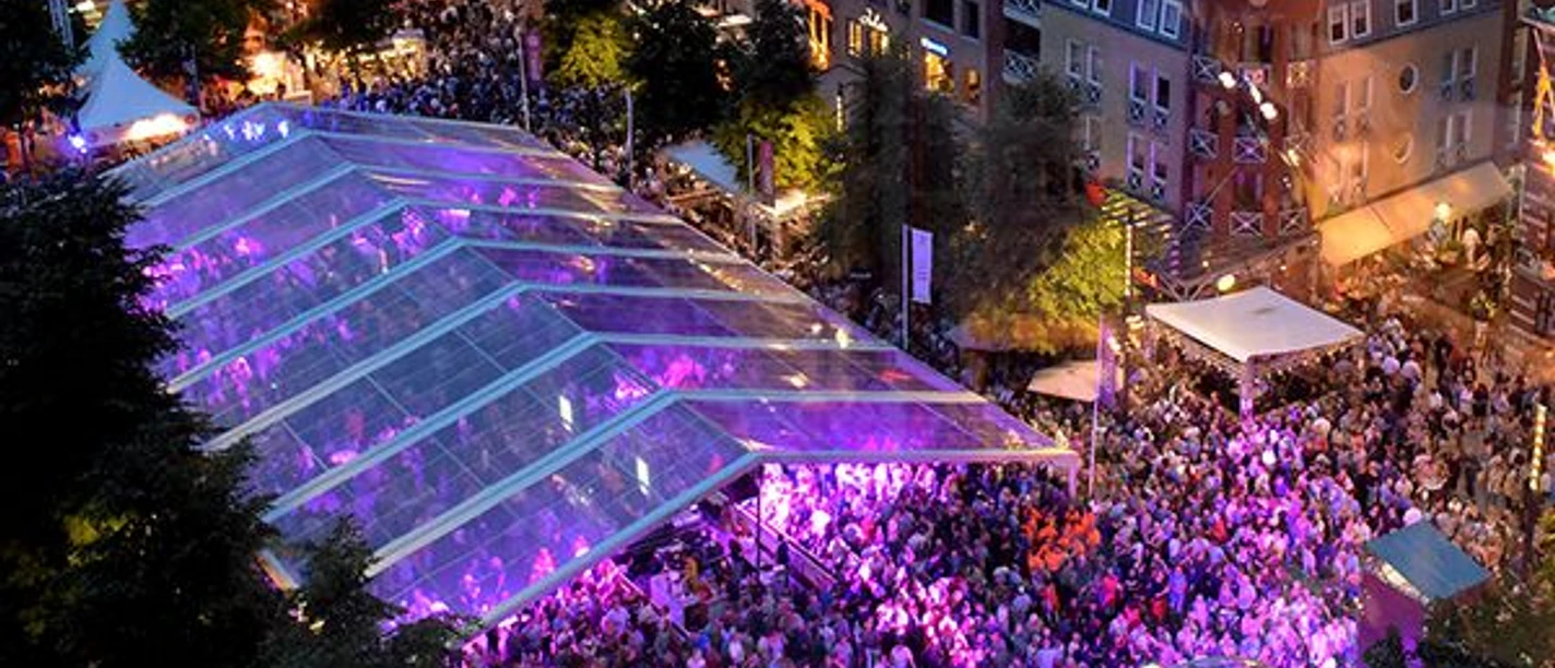 WMC Kerkade (c) WMC.jpg Menschenmenge unter einem transparenten Zelt, das in violettem Licht erstrahlt. Im Hintergrund stehen Gebäude und Bäume.