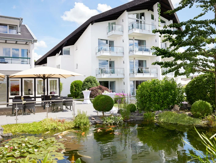 Außenansicht Modernes Hotel mit weißen Balkonen, umgeben von grüner Gartenanlage und Teich mit Seerosen.