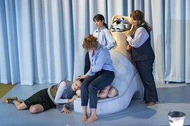 Auf einer Theaterbühne steht ein übergroßer Schuh mit einem Fuß darin. Um den Schuh herum stehen, liegen oder lehnen vier Frauen.