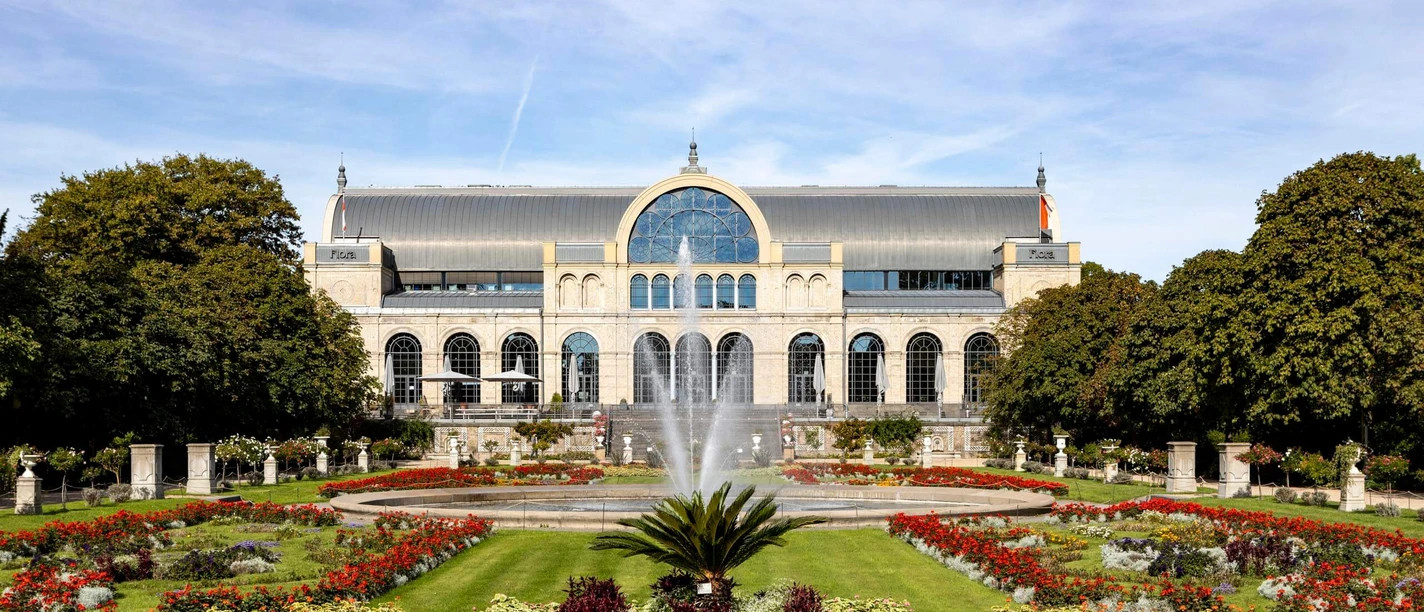 The picture shows the imposing historic façade of the Flora in Cologne, surrounded by flowering gardens and a central fountain. The well-tended park and the symmetrically arranged flowerbeds lend the place a picturesque elegance, while the architecture of the building impresses with its arches and windows.