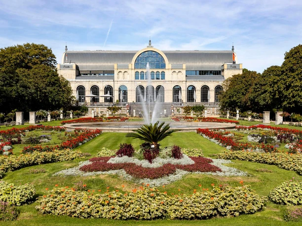 Flora and botanical garden The picture shows the imposing historic façade of the Flora in Cologne, surrounded by flowering gardens and a central fountain. The well-tended park and the symmetrically arranged flowerbeds lend the place a picturesque elegance, while the architecture of the building impresses with its arches and windows.