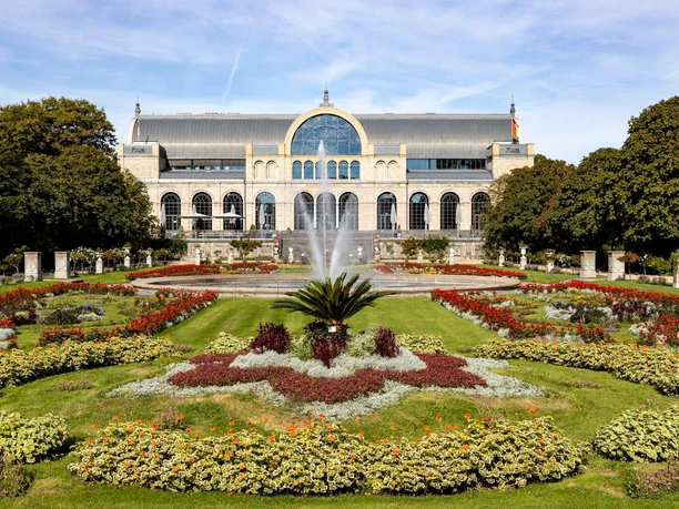 The picture shows the imposing historic façade of the Flora in Cologne, surrounded by flowering gardens and a central fountain. The well-tended park and the symmetrically arranged flowerbeds lend the place a picturesque elegance, while the architecture of the building impresses with its arches and windows.