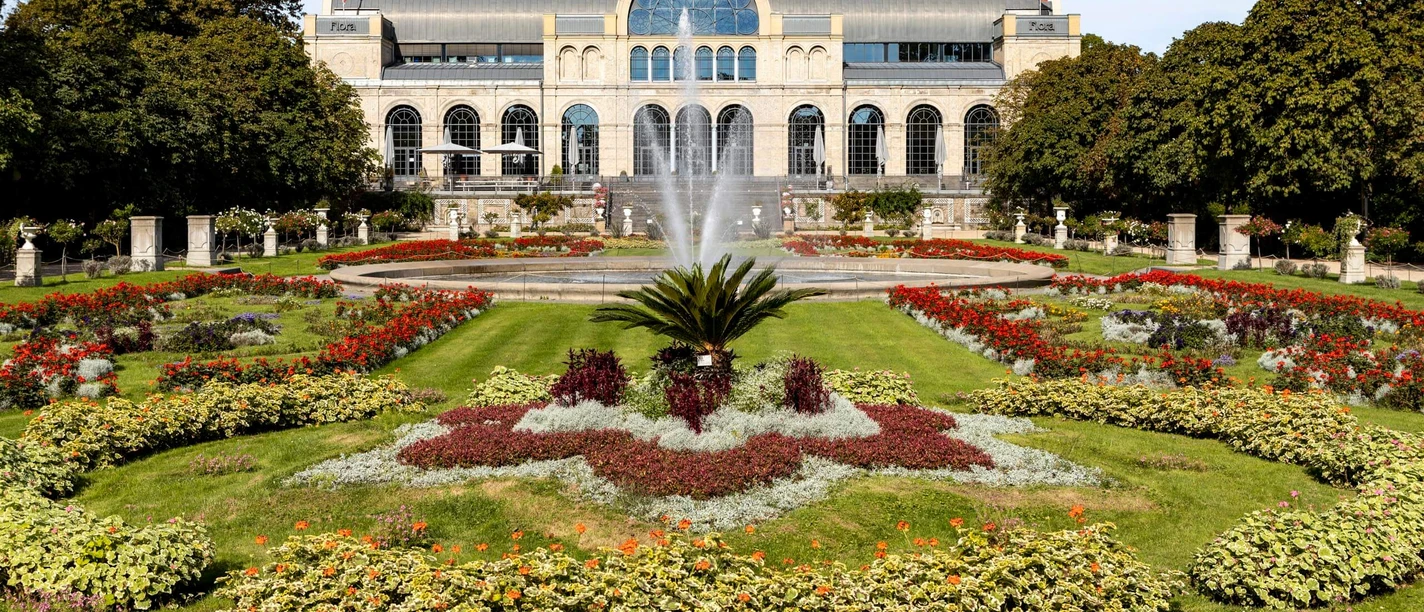 Flora und Botanischer Garten Das Bild zeigt die imposante historische Fassade der Flora in Köln, umrundet von blühenden Gärten und einem zentralen Springbrunnen. Der gepflegte Park und die symmetrisch angelegten Blumenbeete verleihen dem Ort eine malerische Eleganz, während die Architektur des Gebäudes durch ihre Bögen und Fenster beeindruckt.