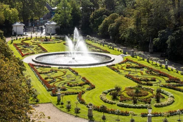 Flora and botanical garden A lush garden with a symmetrical design features a central fountain surrounded by artistically designed flowerbeds and lush greenery. The garden is surrounded by dense trees.