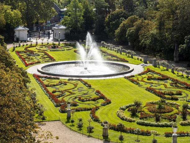 Flora und Botanischer Garten Eine üppige Gartenanlage in symmetrischem Design zeigt einen zentralen Brunnen, umgeben von kunstvoll gestalteten Blumenbeeten, eingebettet in sattes Grün. Der Garten ist von dichtem Baumbestand umrahmt.