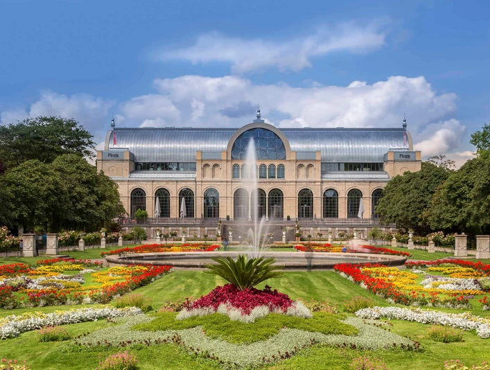 Flora and botanical garden Das Bild zeigt die historische Flora Köln, eine beeindruckende Orangerie mit gläserner Kuppel, eingebettet in farbenprächtige Blumenbeete und einen zentralen Springbrunnen.The picture shows the historic Flora Köln, an impressive orangery with a glass dome, embedded in colorful flowerbeds and a central fountain.