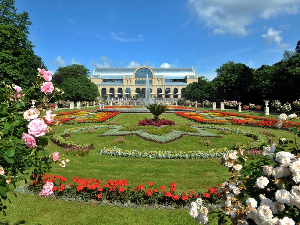 Flora and botanical garden The picture shows the historic Flora in Cologne, surrounded by magnificently blooming gardens, whose symmetrically arranged flowerbeds display a colorful variety. Roses adorn the scenery in the foreground, while the listed building rises majestically into the sky in the background.