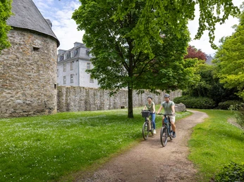Mit dem Rad unterwegs rund um Schloss Hardenberg, Velbert-Neviges Zwei Personen radeln auf einem schmalen Weg entlang einer historischen Schlossmauer im Grünen.