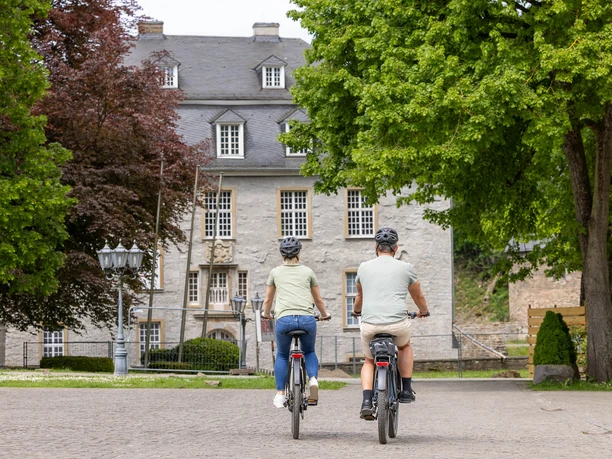 Radtour rund um Schloss Hardenberg, Velbert-Neviges Zwei Radfahrende in Freizeitkleidung radeln vor dem Schloss Hardenberg durch eine von Bäumen gesäumte Allee.