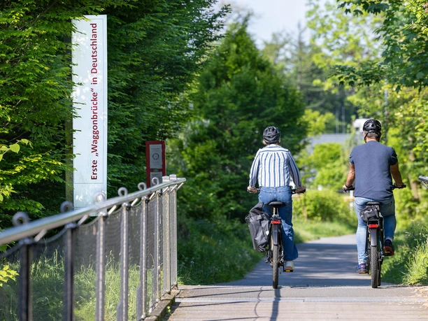 Radtour über die Waggongbrücke in Heiligenhaus Zwei Personen fahren mit Fahrrädern über eine Brücke, umgeben von grüner Vegetation und Sonnenschein.