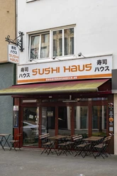 Sushi Haus Die Außenansicht des Sushi Haus zeigt eine große Fensterfront mit geschlossenen Markisen und mehreren Außentischen auf dem Bürgersteig vor dem beige gestrichenen Gebäude.