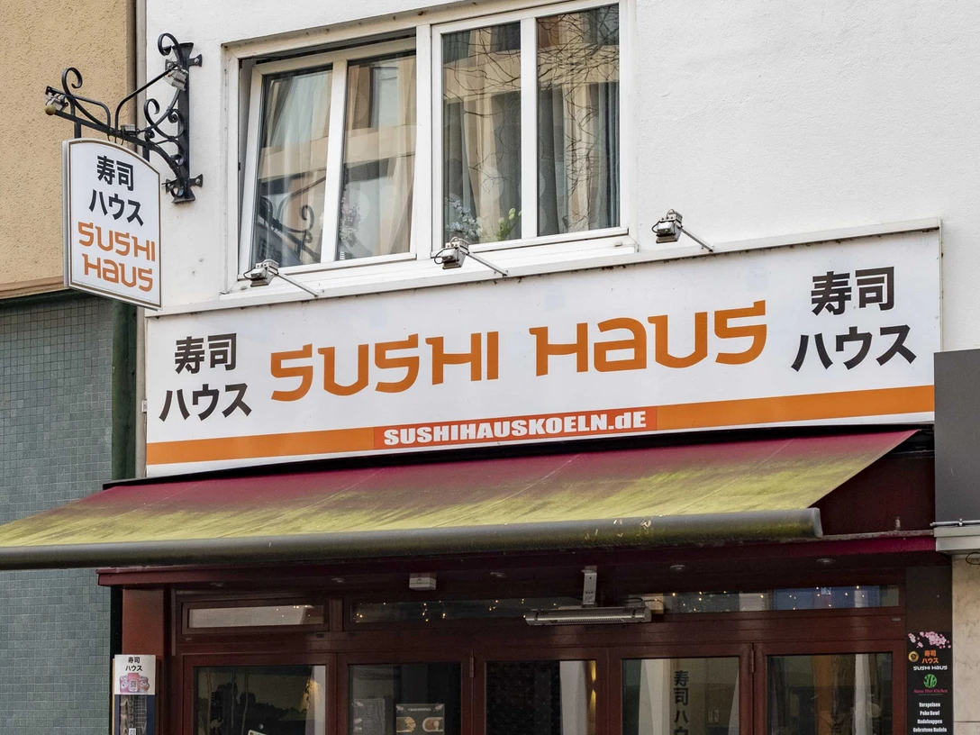 Sushi Haus Die Außenansicht des Sushi Haus zeigt eine große Fensterfront mit geschlossenen Markisen und mehreren Außentischen auf dem Bürgersteig vor dem beige gestrichenen Gebäude. The exterior view of Sushi Haus shows a large window facade with closed awnings and several outdoor tables placed on the sidewalk in front of the beige-painted building.