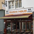 Sushi Haus Die Außenansicht des Sushi Haus zeigt eine große Fensterfront mit geschlossenen Markisen und mehreren Außentischen auf dem Bürgersteig vor dem beige gestrichenen Gebäude. The exterior view of Sushi Haus shows a large window facade with closed awnings and several outdoor tables placed on the sidewalk in front of the beige-painted building.