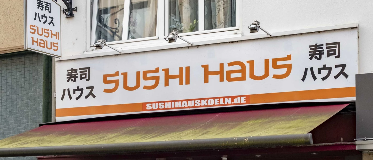 Sushi Haus Die Außenansicht des Sushi Haus zeigt eine große Fensterfront mit geschlossenen Markisen und mehreren Außentischen auf dem Bürgersteig vor dem beige gestrichenen Gebäude.