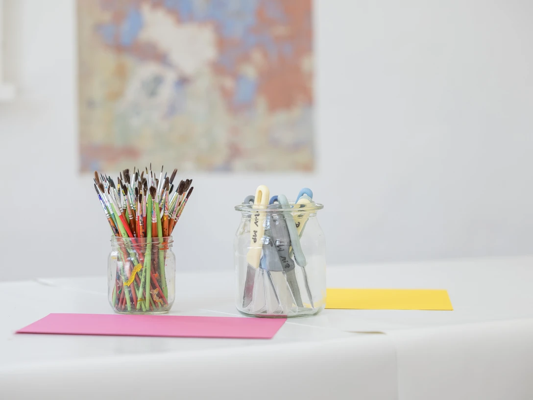 Überraschungswerkstatt_Philipp Harms.jpg Malutensilien in Gläsern stehen auf einem weißen Tisch, ein Kunstwerk ist im Hintergrund sichtbar.