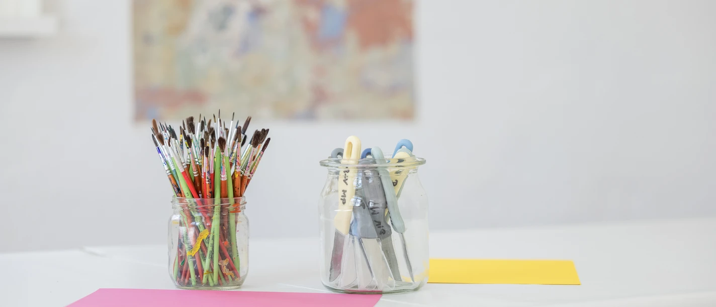 Überraschungswerkstatt_Philipp Harms.jpg Malutensilien in Gläsern stehen auf einem weißen Tisch, ein Kunstwerk ist im Hintergrund sichtbar.