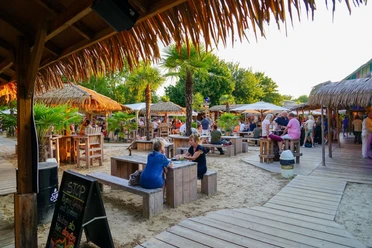 Aloha-Beachclub Herford, L Schneider (2).jpeg Strandbar mit Holzmöbeln und Palmen, Menschen genießen Getränke bei Sonnenuntergang in tropischem Flair.