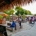 Aloha-Beachclub Herford, L Schneider (2).jpeg Strandbar mit Holzmöbeln und Palmen, Menschen genießen Getränke bei Sonnenuntergang in tropischem Flair.